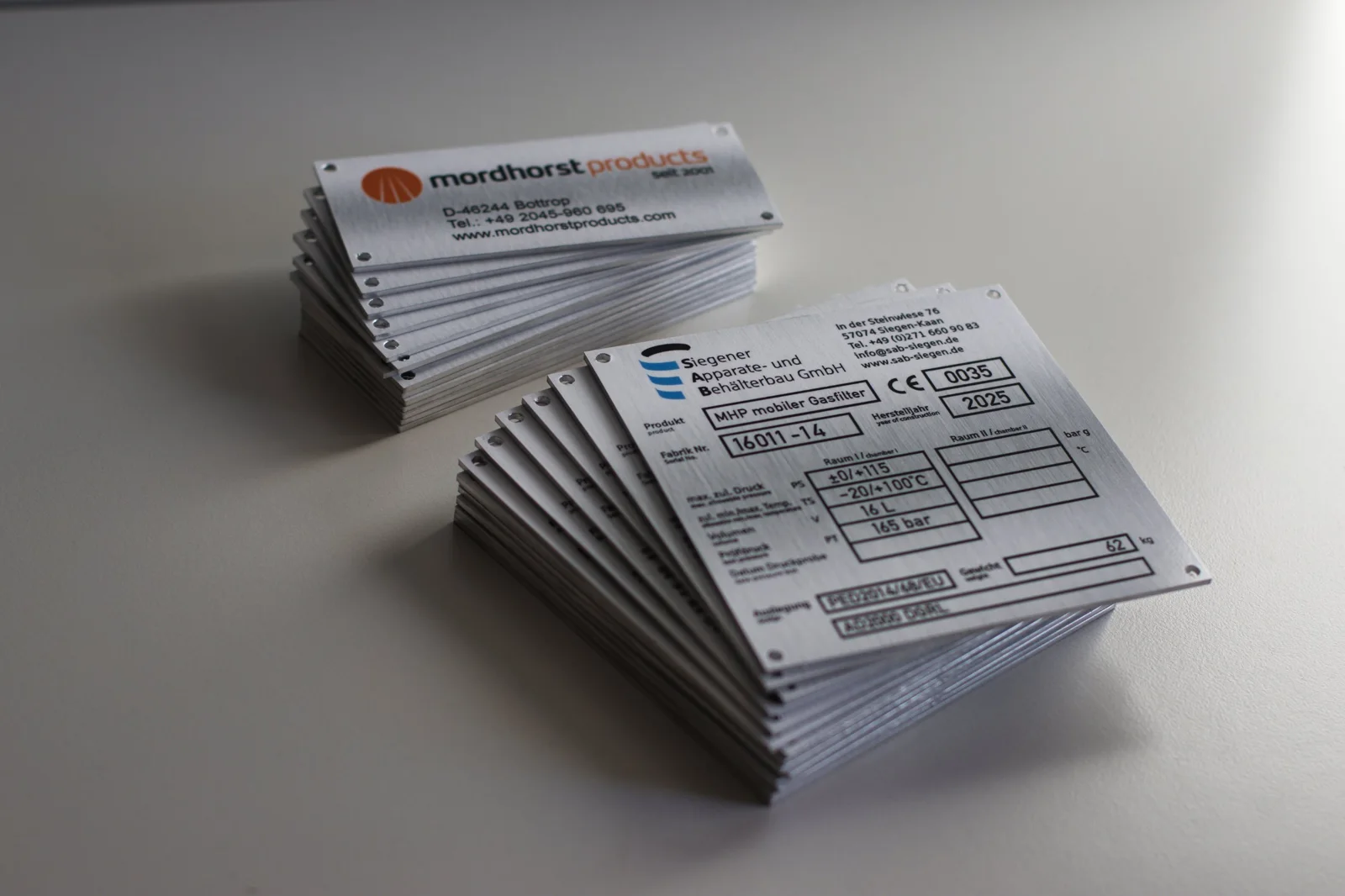 Typenschilder aus gebürstetem Aluminium für SAB Siegen mit CE-Kennzeichnung und Datenfeldern, dahinter Firmenschilder für mordhorst products